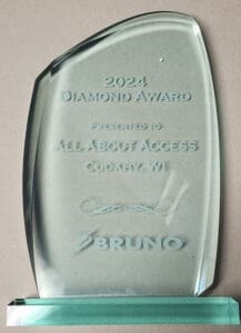 A glass award with engraved text reading 2024 Diamond Award Presented to All About Access Cudahy, WI above the logo and name Bruno. The award is standing on a green glass base.