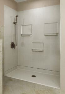 A modern walk-in shower with light-colored tiles, three built-in shelves, a dark bronze showerhead, and a single handle. The shower has a low, white threshold and tiled floor outside.