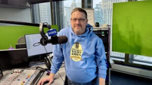 A man wearing glasses and a blue Chicagos Very Own hoodie stands in a radio studio beside a mixing console and a WGN microphone, with city buildings visible through the window behind him.