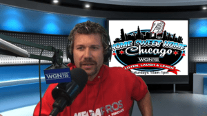 A man in a red hoodie speaks into a microphone in a radio studio. Behind him is a screen displaying the Home Sweet Home Chicago logo with show details and a WGN Radio 720 sign on the microphone.