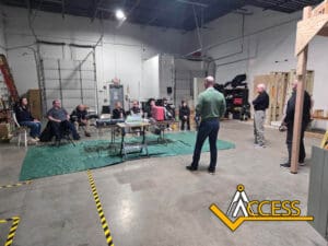 A group of people sit and stand around tables in a large industrial warehouse, listening to a speaker. The ACCESS logo is visible in the lower right corner of the image.