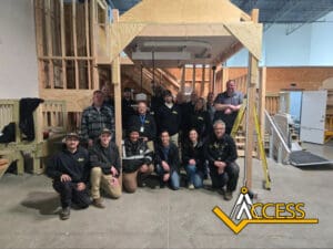 A group of 15 people pose and smile inside a warehouse with wooden structures and stairs. Most wear casual clothing and hats. The Access logo appears in the lower right corner.