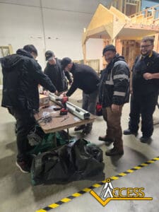 Five people work together on a project at a table in a workshop, assembling materials. Wooden structures and tools are visible, with the Access logo in the bottom right corner.