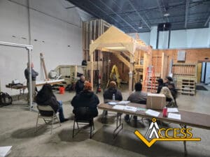 A group of people sits at tables in a warehouse classroom, listening to an instructor near a partially built wooden structure. Ladders and building materials are visible, with the ACCESS logo in the corner.