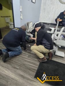 Three men kneel on a wood floor while installing or repairing a stair lift in an indoor setting. The ACCESS company logo is visible in the bottom right corner of the image.