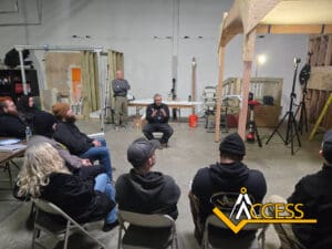 A man sits and speaks to a group of people seated in a workshop or training area, with construction materials and equipment around. A logo reading ACCESS is displayed in the lower right corner.