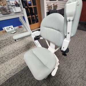 A light gray stair lift chair with armrests and seatbelt is positioned at the base of a carpeted staircase in a showroom with display shelves and other mobility products in the background.