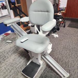 A stairlift with a light gray cushioned seat and armrests is installed on a carpeted staircase in a showroom, with mobility aids and informational posters visible in the background.