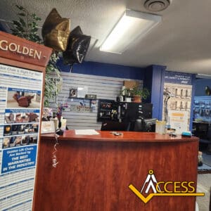 Reception area with a wooden desk, balloons, office supplies, and informational brochures. A sign reads GOLDEN and the ACCESS logo is visible. The background shows shelves with products and a printer.