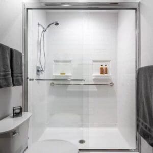 The Walk-In Shower by Best Bath features modern white tiles, sliding glass doors, a silver showerhead, grab bar, built-in shelves for toiletries, and nearby dark gray towels. A portion of a toilet appears in the foreground.