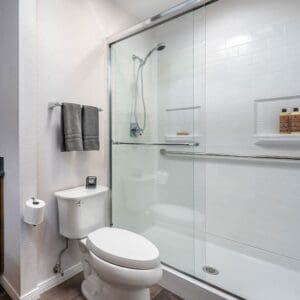 Modern bathroom with a Walk-In Shower by Best Bath featuring white subway tile, ADA compliance, glass enclosure, wall-mounted showerhead, built-in shelf with toiletries, white toilet, toilet paper holder, and two gray towels on a metal rack.