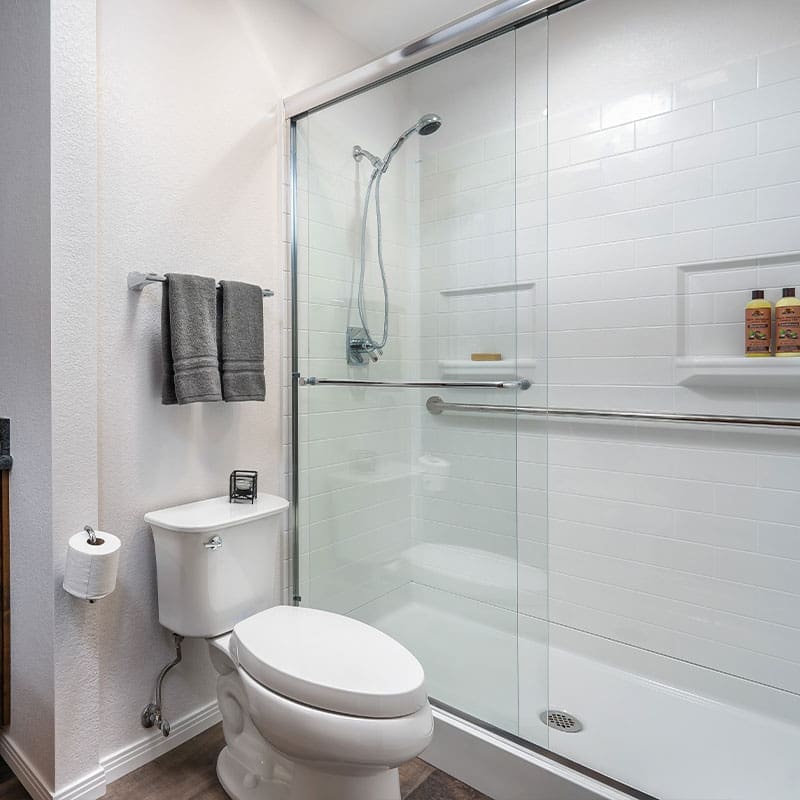 Modern bathroom with a Walk-In Shower by Best Bath featuring white subway tile, ADA compliance, glass enclosure, wall-mounted showerhead, built-in shelf with toiletries, white toilet, toilet paper holder, and two gray towels on a metal rack. Modern bathroom with a Walk-In Shower by Best Bath featuring white subway tile, ADA compliance, glass enclosure, wall-mounted showerhead, built-in shelf with toiletries, white toilet, toilet paper holder, and two gray towels on a metal rack.