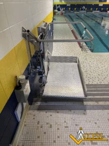 A platform lift is installed next to an indoor swimming pool, providing accessible entry from the tiled pool deck down to the water. The Access logo is in the lower right corner.