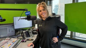 A woman in a black hoodie stands and smiles in a radio station studio, with a microphone labeled WGN and a soundboard in front of her. Large windows show city buildings in the background.