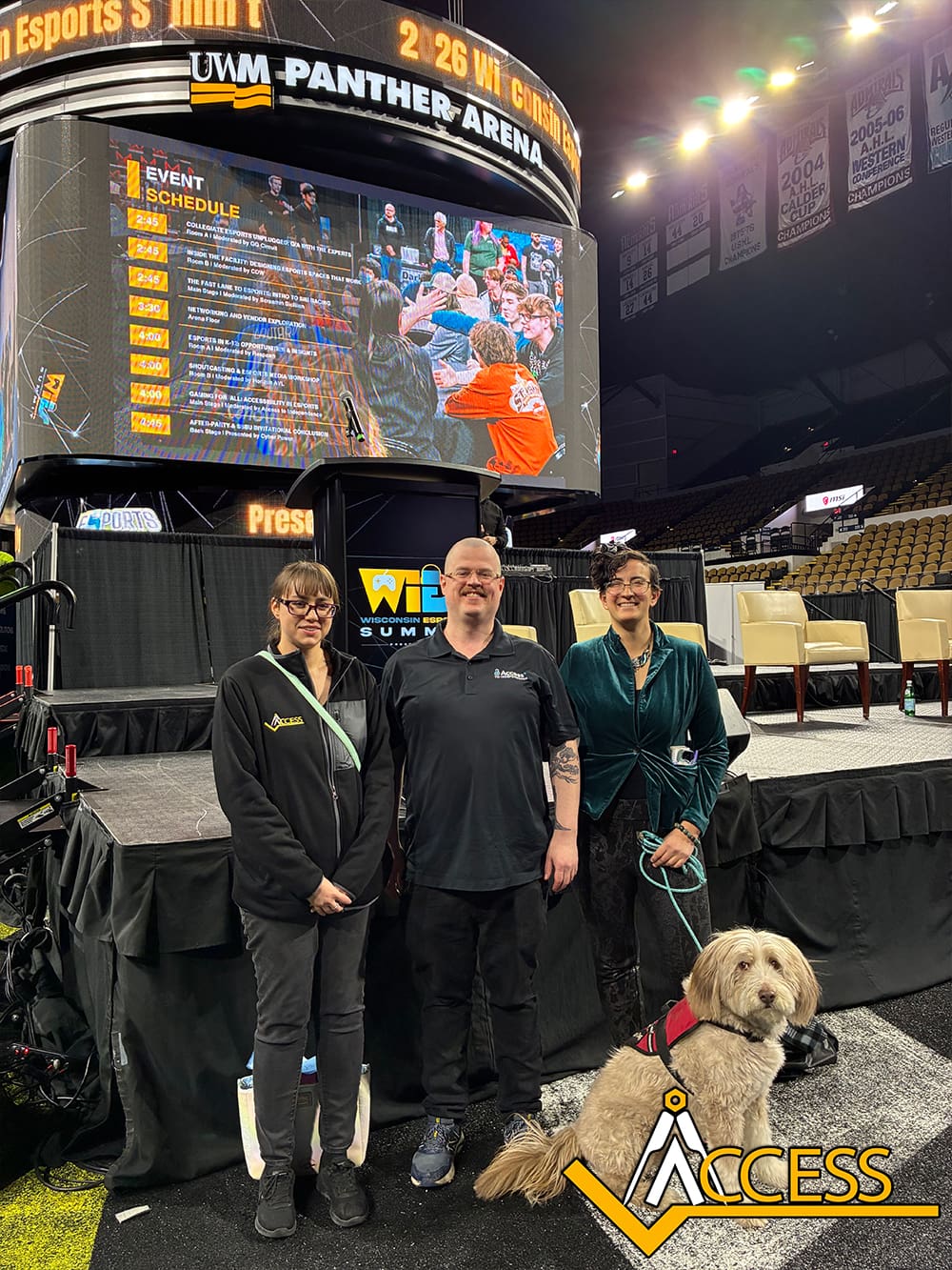 Three people and a fluffy dog pose in front of a large screen displaying an event schedule and crowd at UWM Panther Arena. The ACCESS logo is at the bottom right of the image.