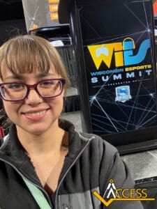 A woman with glasses smiles for a selfie in front of a podium displaying the Wisconsin Esports Summit logo and graphics. The ACCESS logo is visible in the lower right corner.