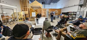 A group of people sit in chairs facing two instructors presenting blueprints in an industrial workshop with wood structures and shelves; the Access logo appears in the lower right corner.