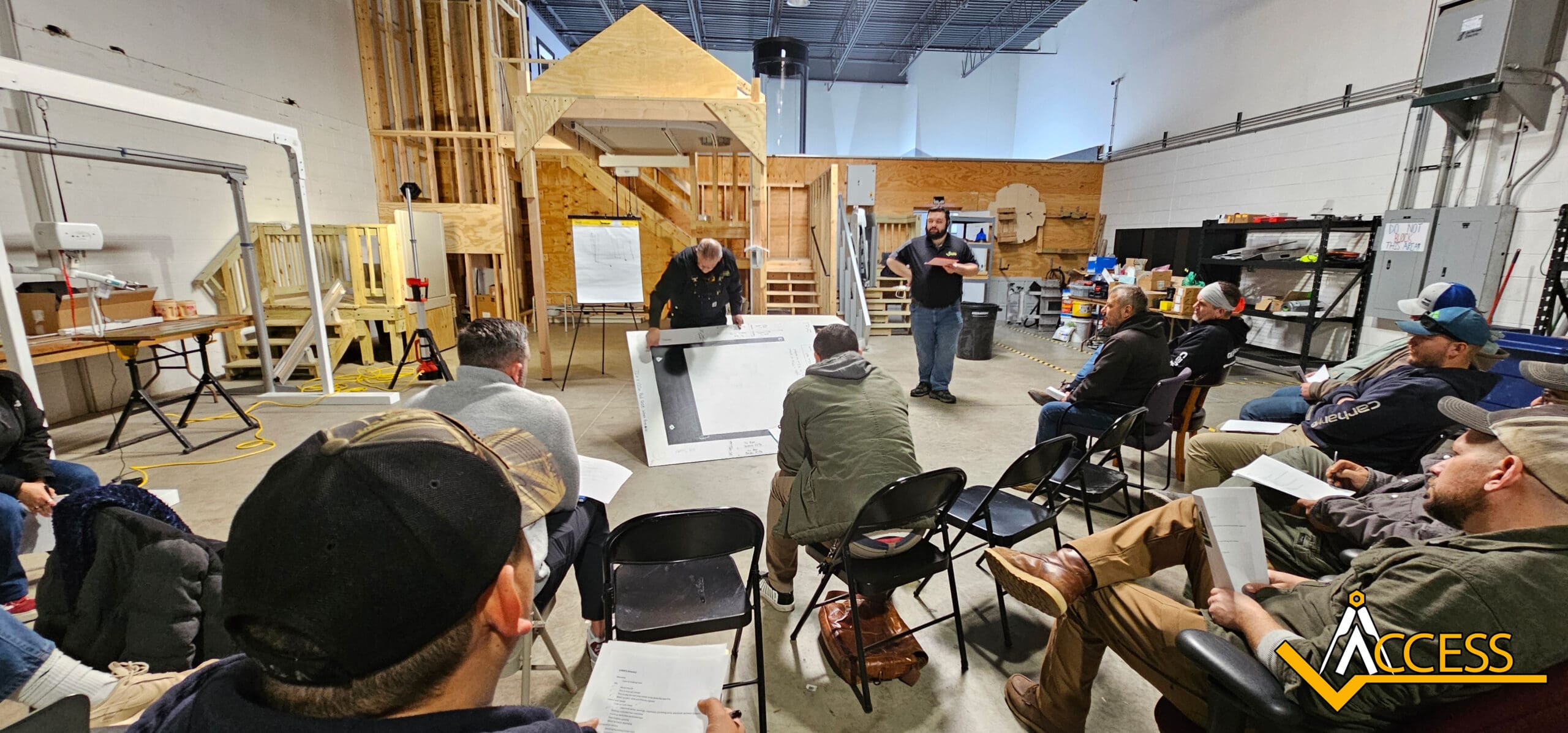 A group of people sit in chairs facing two instructors in a workshop setting, with construction models and materials visible in the background. One instructor points to a large blueprint. The Access logo is in the bottom right corner.