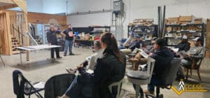 A group of people sit in chairs inside a workshop, listening to two men standing at the front near a table. Shelves of supplies and tools are visible in the background. A logo reading ACCESS appears in the lower right corner.