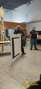 A man holds a large board covered in tape and writing in a workshop setting, while another man stands with arms crossed in the background. Workshop materials and tools are visible around them. ACCESS logo is in the bottom right corner.