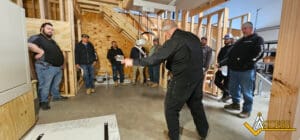 A man in black coveralls demonstrates equipment to a group of seven men standing in a partially constructed wooden room. The ACCESS logo is visible in the bottom right corner.
