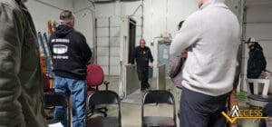 A man is walking out of a secure metal entryway in a warehouse, while several people stand and watch. Empty chairs are in the foreground, and the Access company logo appears in the bottom right corner.