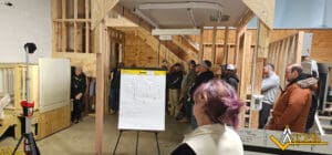 A group of people stand in a workshop, listening to a presenter near a whiteboard with diagrams. Wooden framing, stairs, and various equipment fill the space. The Access logo is visible in the bottom right corner.