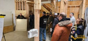 A group of people stands in a workshop, listening to an instructor demonstrating equipment. The setting features wooden frames and tools, with an Access logo visible in the bottom right corner.