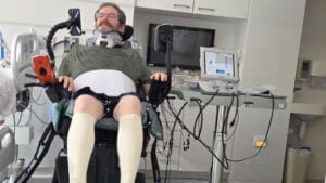A man wearing a neck brace and medical stockings sits in a hospital chair surrounded by medical equipment and monitors in a clinical room.