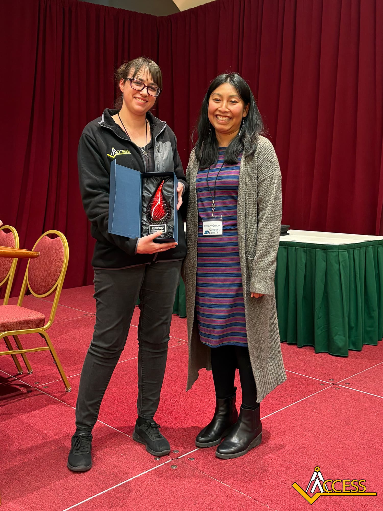 Two women stand on a red carpeted stage with red curtains; one holds a red glass award in a blue case. Both are smiling. On the bottom right is a logo that reads ACCESS.