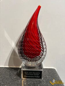 A glass award shaped like a teardrop with red and black swirls, sitting on a black base. The nameplate reads: 2026 John Nousaine Relentless Badger Cassandra Potter. The ACCESS logo is in the corner.