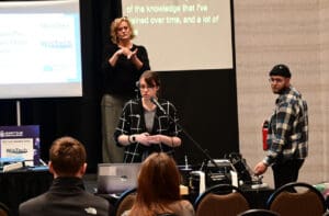 A woman stands at a podium speaking to an audience, with a laptop in front of her. Behind, a sign language interpreter translates, while a man stands nearby holding a water bottle. Presentation slides are visible to the side.