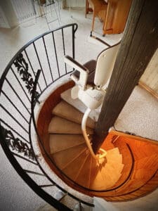A spiral staircase with carpeted steps and a black metal railing features a stair lift chair attached to the inner curve, allowing accessibility between floors in a cozy, warmly lit home interior.