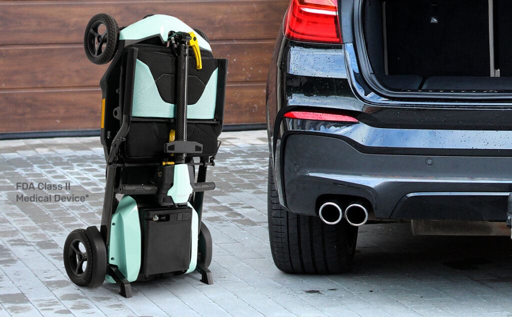 A folded teal and black Go Go Super Portable 4-Wheel Scooter by Pride Mobility stands upright on a driveway beside the open trunk of a dark car. A wall label reads “FDA Class II Medical Device.”.