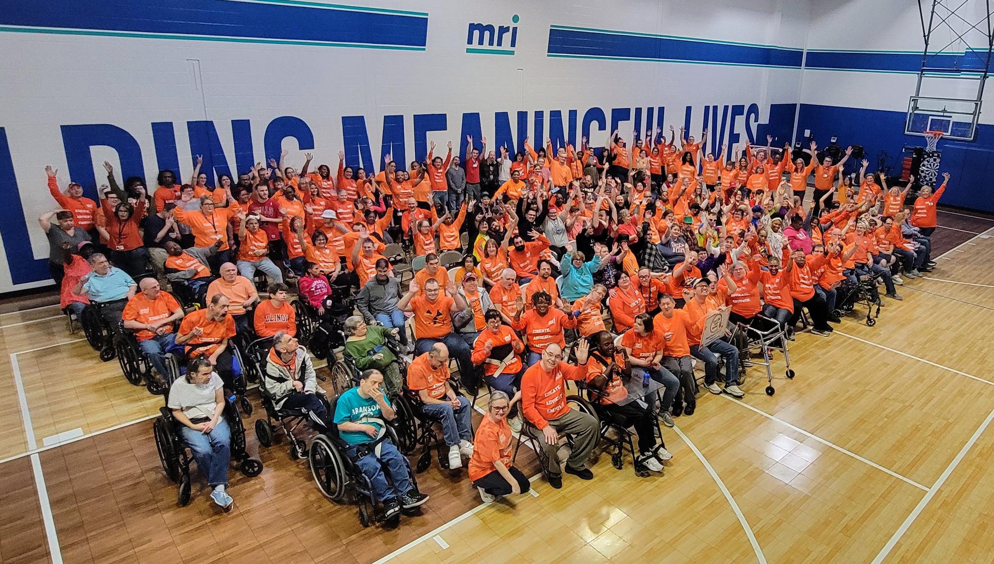 A large, diverse group of people, many in wheelchairs, wear matching orange shirts and gather in a gymnasium, smiling and raising their arms in celebration. The words MEANINGFUL LIVES are visible on the wall behind them.