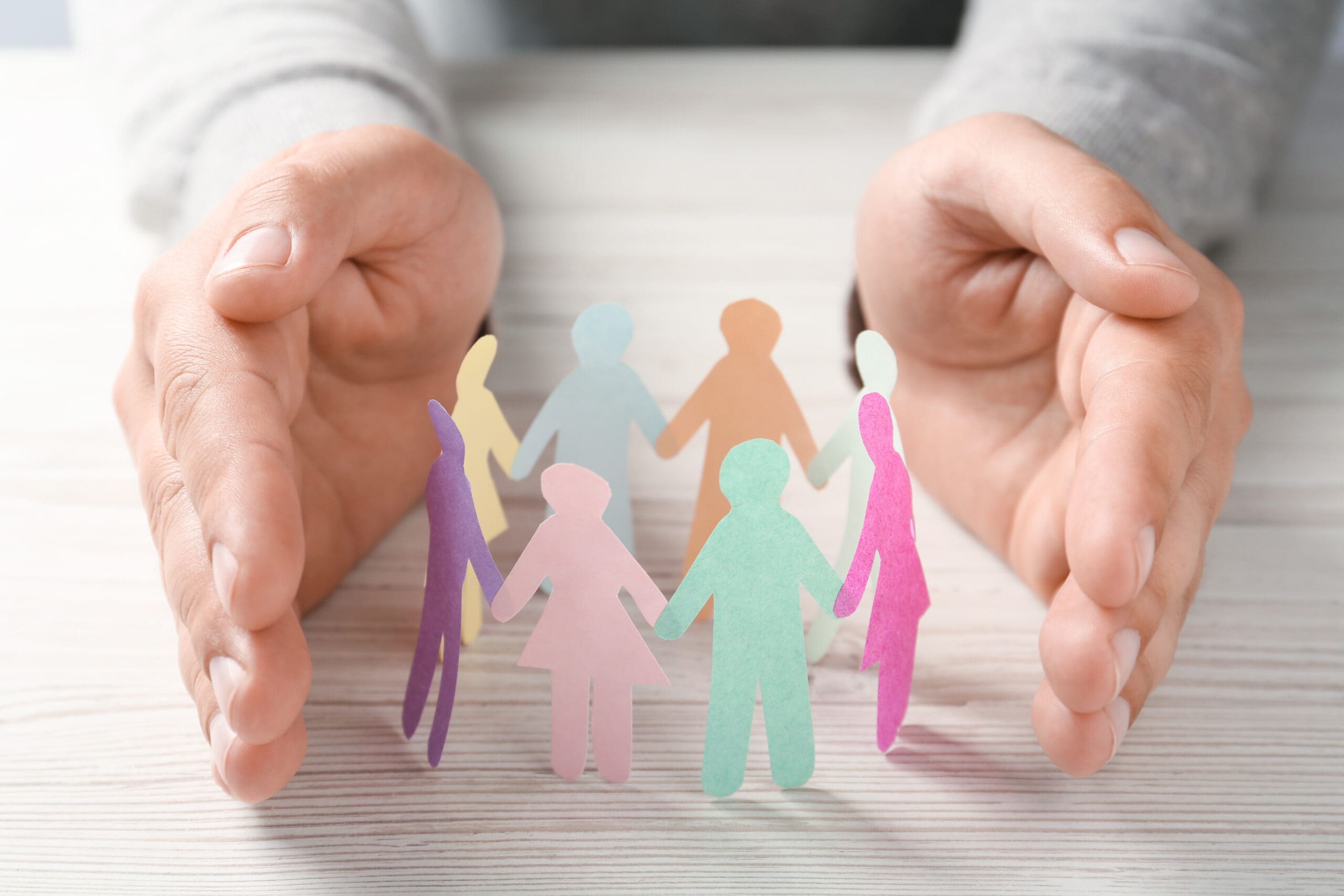 Two hands gently encircle a group of colorful paper cutout figures, symbolizing unity, support, and protection on a light wooden surface.