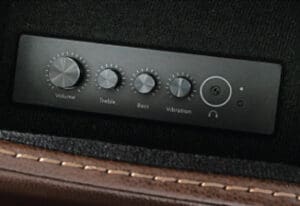 A close-up of a black control panel on the VivaLift Premier PLR-5900 Lift Chair by Pride Mobility, featuring four dials (volume, treble, bass, vibration), a power button, headphone jack, and indicator lights set against dark leather with brown trim.