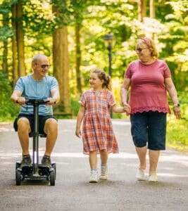 An elderly man rides a Go Go Endurance AL+-Wheel Scooter by Pride Mobility, smiling as he holds hands with a young girl and an elderly woman, all enjoying a sunny walk together on a wooded path.