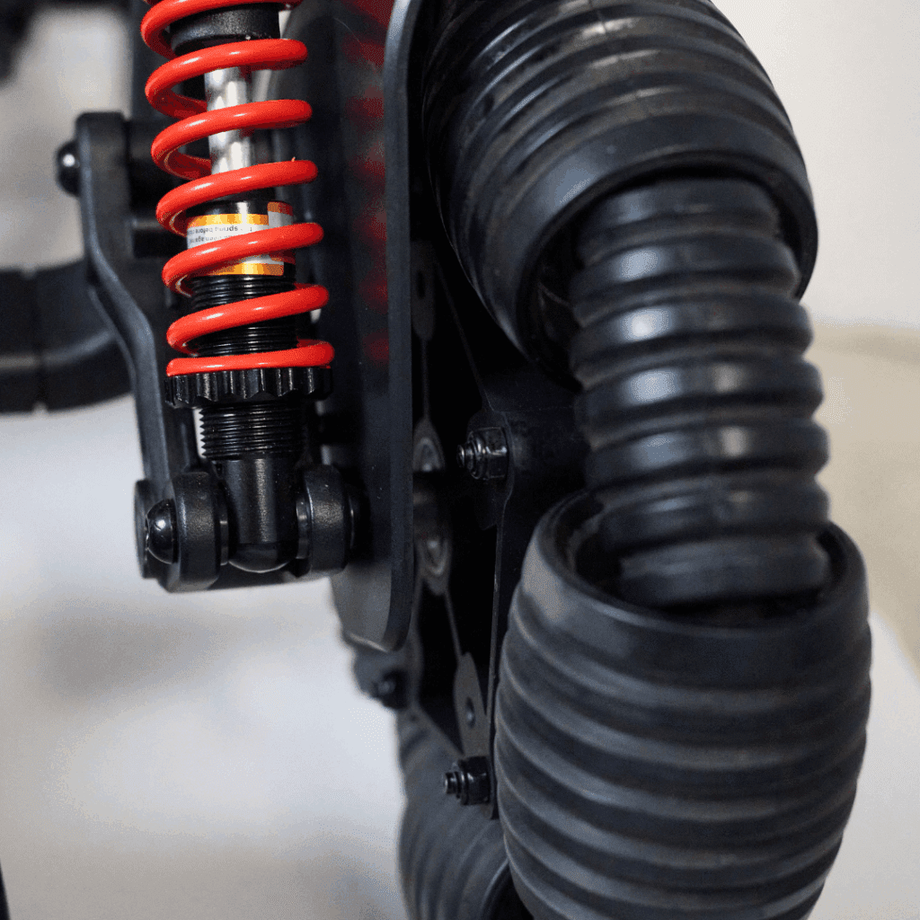 Close-up of the suspension system on the Ally Pro - Power Wheelchair by Golden, featuring a red coil spring and large black ribbed hoses with detailed parts for advanced mobility.