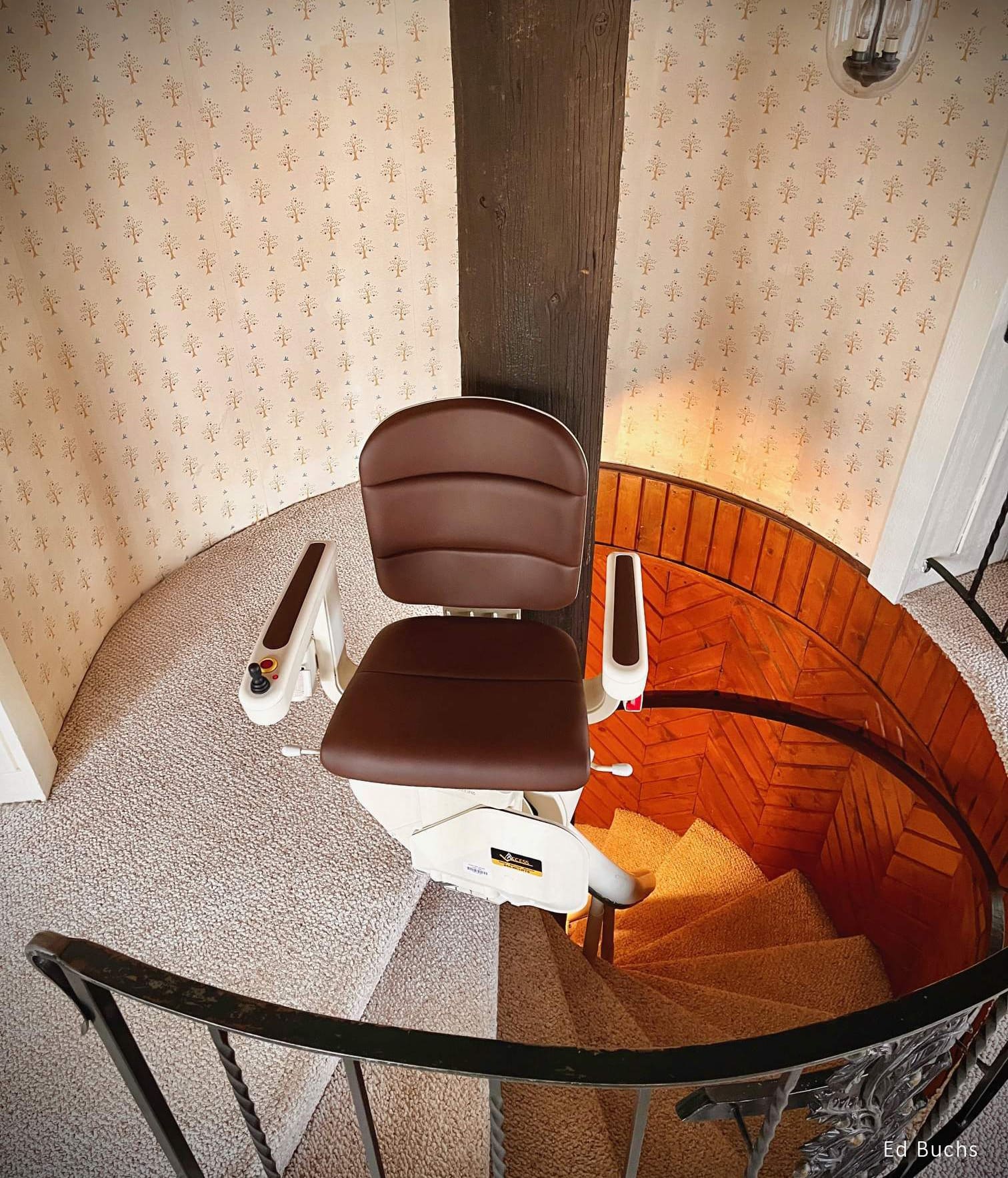 A brown Savaria stair lift chair is installed on a spiral staircase with carpeted steps, wrapping around a central wooden pole in a vintage-style interior with patterned wallpaper.