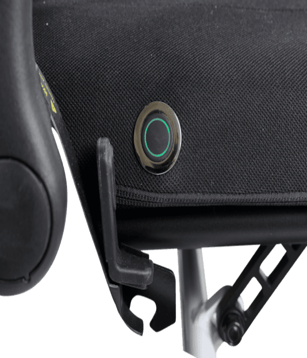 Close-up of a black fabric seat on the Ally - Power Wheelchair by Golden, showing a round button with a green ring light and an attached plastic clip under the seat.