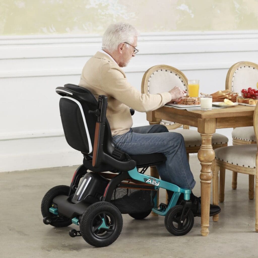 An older man sits at a wooden dining table in his Ally - Power Wheelchair by Golden, enjoying breakfast with orange juice, toast, and fruit against a light-colored indoor wall.