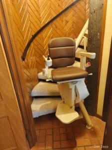 A brown Savaria stair lift chair is installed on a carpeted staircase with wooden paneled walls and a tiled floor at the base. The chair faces forward, ready for use.