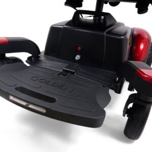 Close-up of a black footrest platform with "Golden" embossed, attached to the red and black BuzzAbout - Power Wheelchair by Golden, highlighting the wheels and frame.