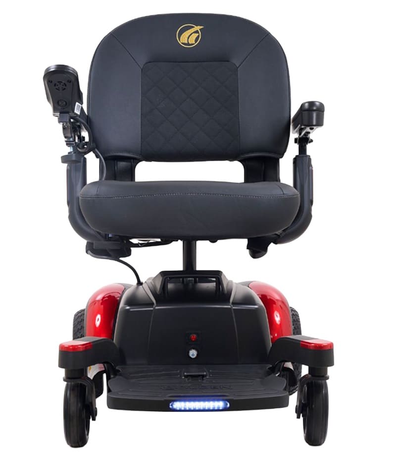 A front view of the BuzzAbout - Power Wheelchair by Golden in black and red, featuring a padded seat, armrests, left-side joystick control, and a small LED light on the lower front. A front view of the BuzzAbout - Power Wheelchair by Golden in black and red, featuring a padded seat, armrests, left-side joystick control, and a small LED light on the lower front.