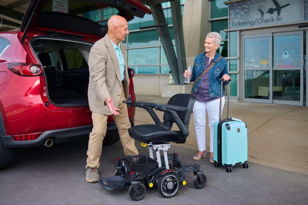 A man and woman smile at each other outside an airport. The man stands by a red car with its trunk open, holding a Buzzaround CarryOn - Power Wheelchair by Golden, while the woman stands nearby with a teal suitcase and water bottle.