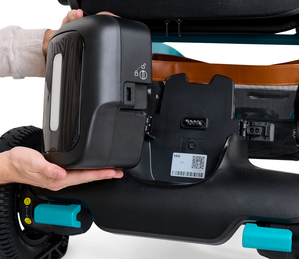 Close-up of a person installing or removing a black battery pack from the Ally - Power Wheelchair by Golden, with visible wheels and a QR code label on the frame.