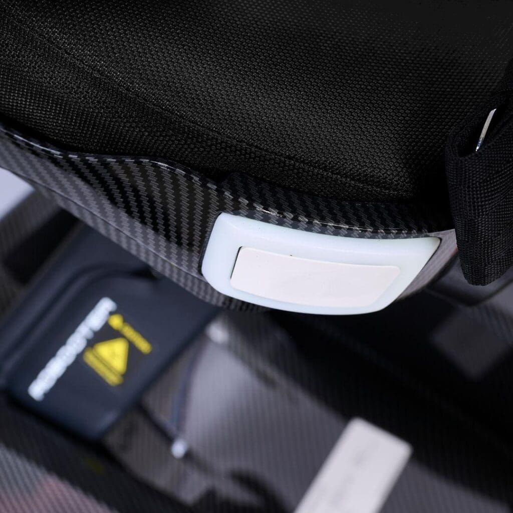 Close-up of a car seat buckle with a white release button on a black, carbon fiber-textured seat—perfect for securing your Ally Sport - Power Wheelchair by Golden. A partially visible warning label appears in the background.