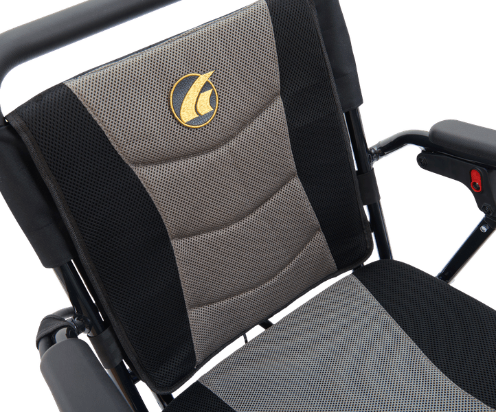 Close-up of the Stride - Power Wheelchair by Golden, showing its black and gray textured seat fabric, gold emblem on the backrest, visible armrests, and part of the lightweight power wheelchair frame.