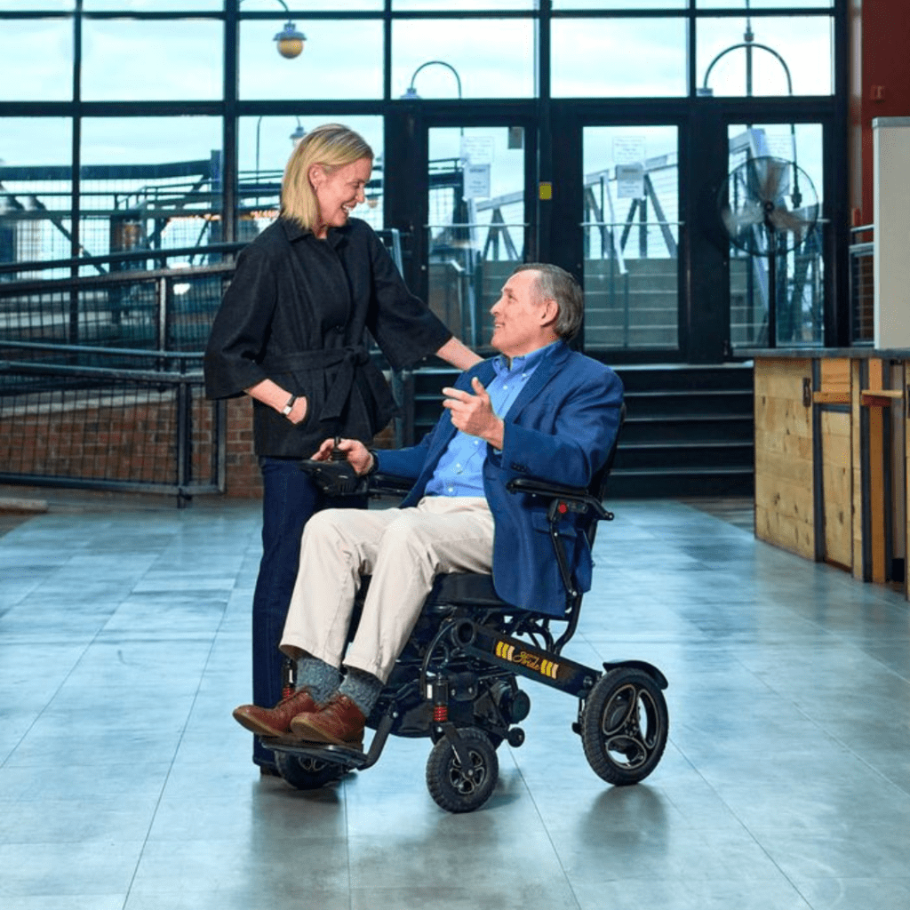 A woman smiles and talks with a man in a blue jacket who is sitting indoors near glass doors in a Stride - Power Wheelchair by Golden.